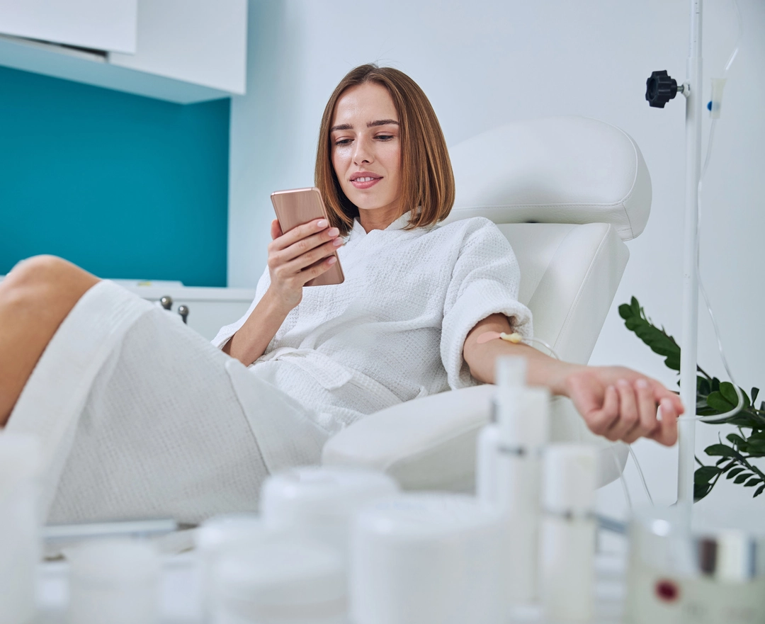 A woman in a white bathrobe relaxes in a spa chair getting IV infusion, holding a smartphone, surrounded by skincare products and a green plant A woman in a white bathrobe relaxes in a spa chair getting IV infusion, holding a smartphone, surrounded by skincare products and a green plant